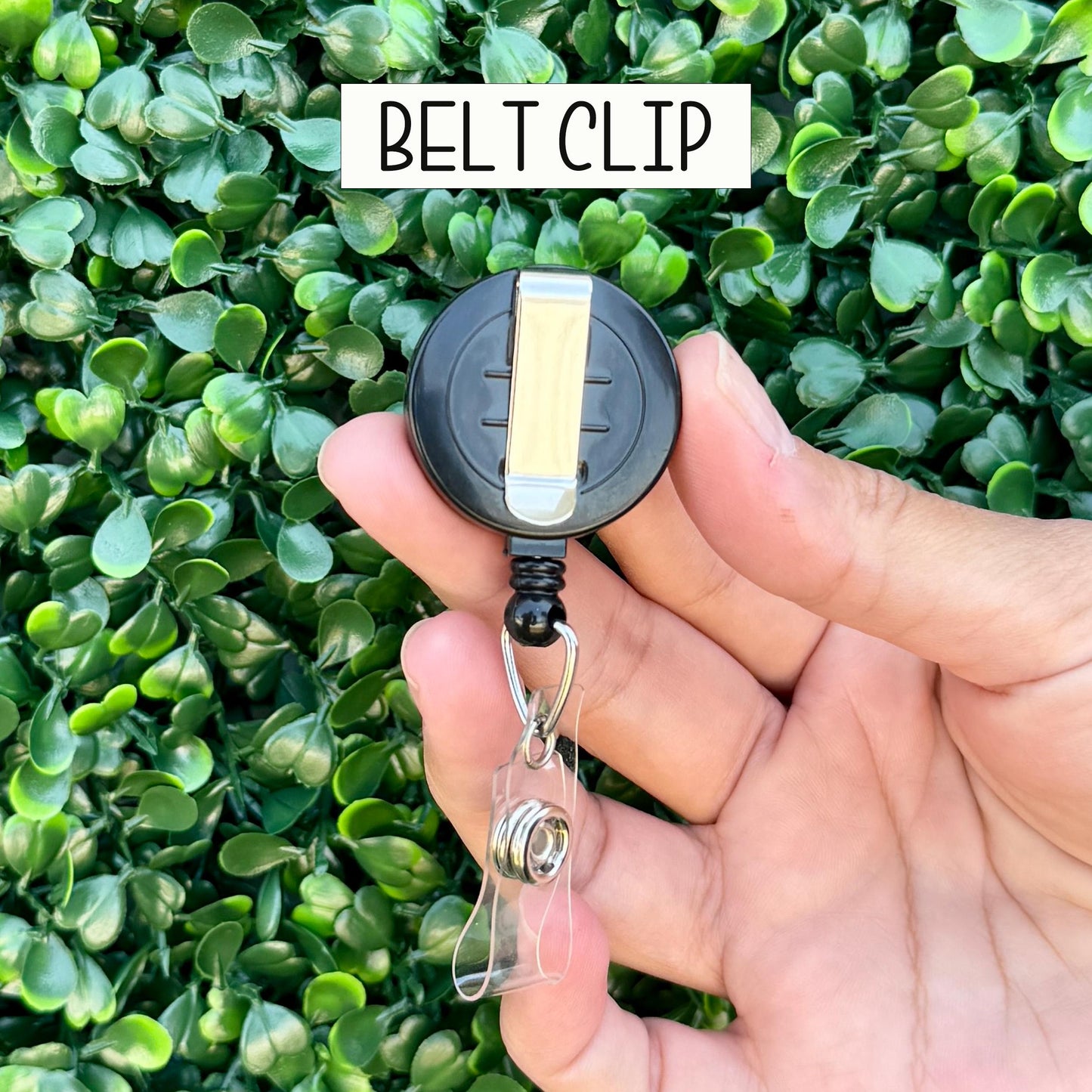 A hand holding a black belt clip with a clear plastic card holder attached to it, against a background of green leaves.