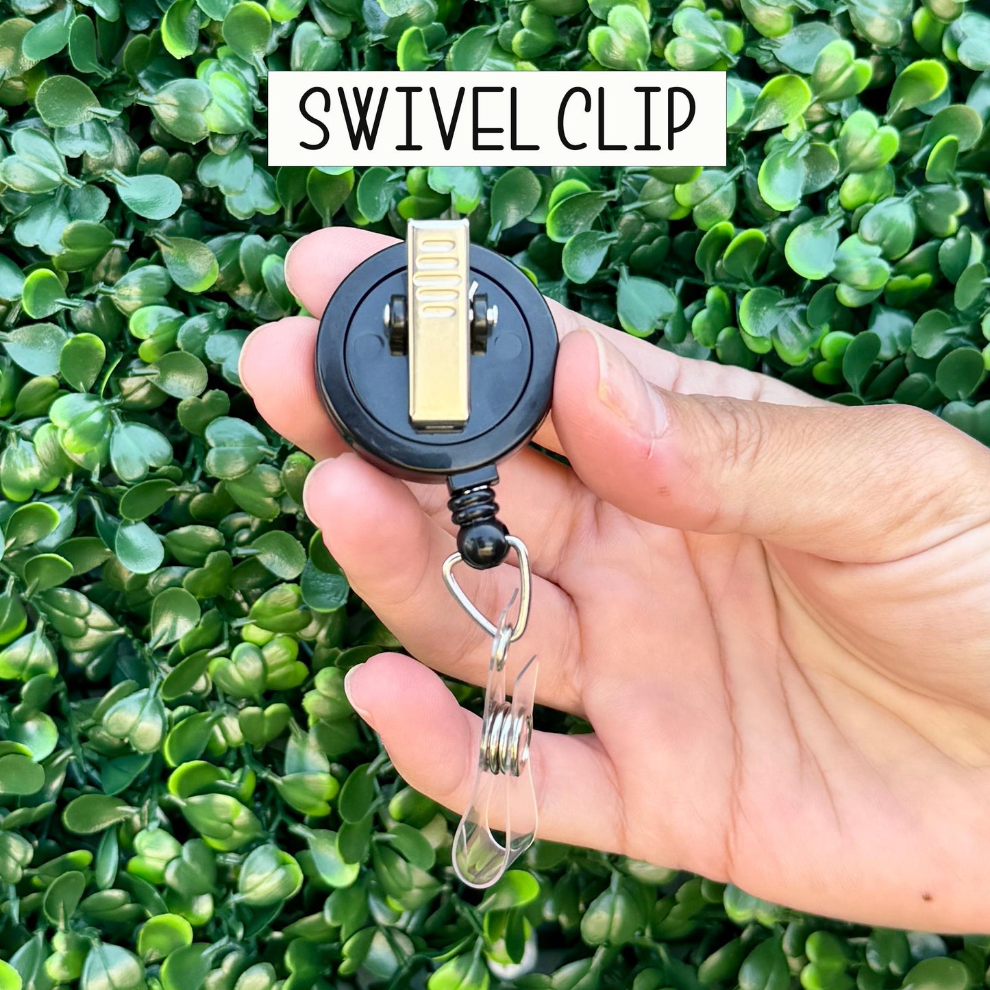 A hand holding a black and gold Swivel Clip with a clear plastic loop attached to it, against a background of green leaves.